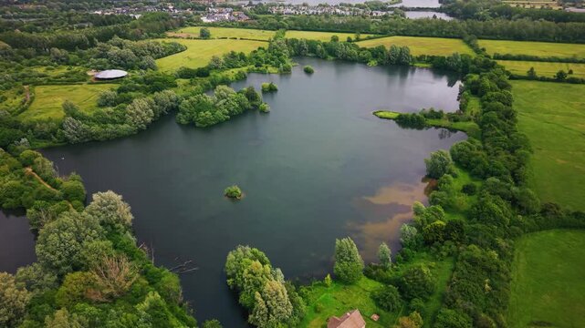 Drone aerial footage showing a large freshwater lake bordered by woodland, reed beds, wetlands and open green countryside. The scene captures a peaceful natural landscape with islands, trees
