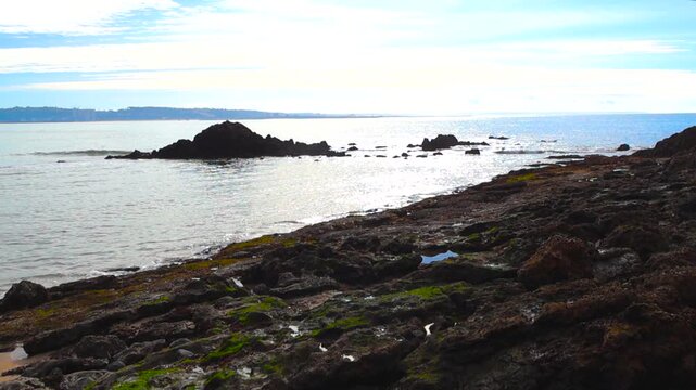 Sunny view of the Tagus river estuary from a rocky coastline covered in moss, with calm water gently lapping the shore under a bright sky with some clouds and the city in the background