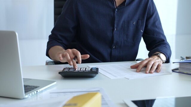 Businessman in a blue shirt examining financial documents at an office desk, using a calculator amidst paperwork and folders. Audit and taxes, business concept