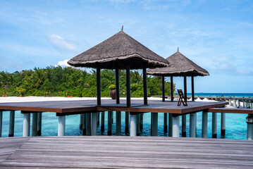 Fototapeta premium Overwater deck with thatched pavilions extends above turquoise lagoon in the Maldives, Male Atoll. Empty lounge chairs sit on clean wooden planks under bright daylight and clear sky.