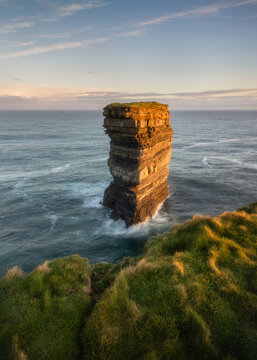 View of Dun Briste Sea Stack with its layered rock formations standing in the Atlantic Ocean near grassy cliffs during golden hour in County Mayo, Ireland.