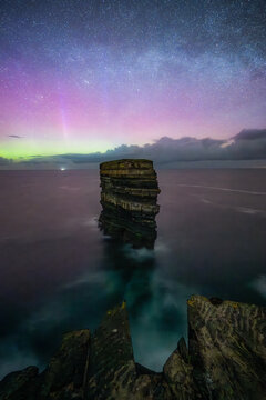 View of Dun Briste Sea Stack under the Aurora Borealis and a starry night sky with dark ocean waves and rocky cliffs in County Mayo, Ireland.