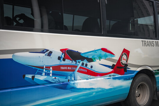 A shuttle bus shows a Trans Maldivian Airways seaplane graphic and TMA logo in Male, Maldives. Bright branding and sharp graphics appear under diffuse daylight.
