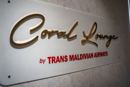 A polished white plaque with gold script and red lettering reads Coral Lounge by Trans Maldivian Airways at Velana International Airport in Male, Maldives, under soft indoor light.
