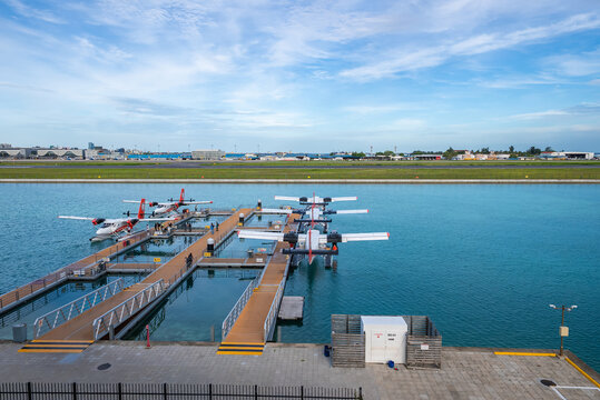 Twin Otter floatplanes dock at wooden piers on turquoise water in Male, Maldives, with a lagoon runway beyond. Daylight, wide angle view shows orderly staging and calm water.