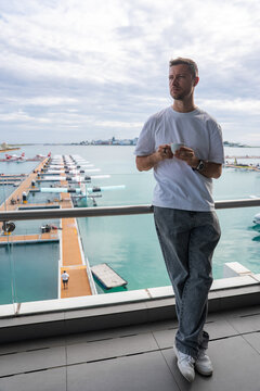 A man stands on a balcony facing the TMA seaplane terminal near Velana in Male, Maldives. Moored seaplanes, orange walkways, and an overcast sky frame the harbor.