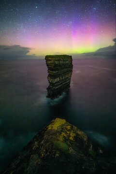 View of the Dun Briste Sea Stack from a rocky cliff under a starry night sky illuminated by the vibrant colors of the Aurora Borealis over the Atlantic Ocean in County Mayo, Ireland.