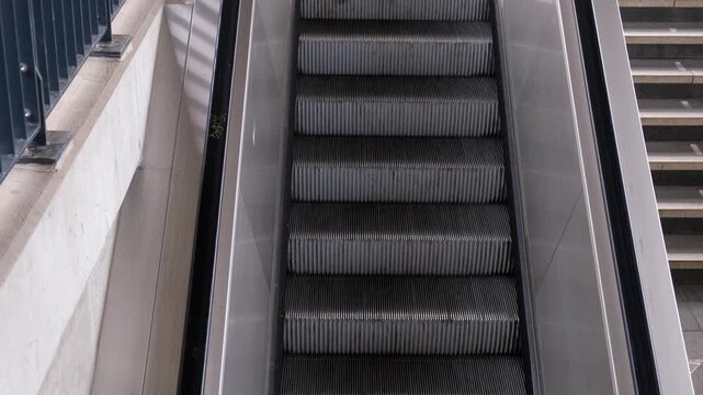A top-down view of a functional silver metal escalator moving downwards next to standard concrete stairs at a station.