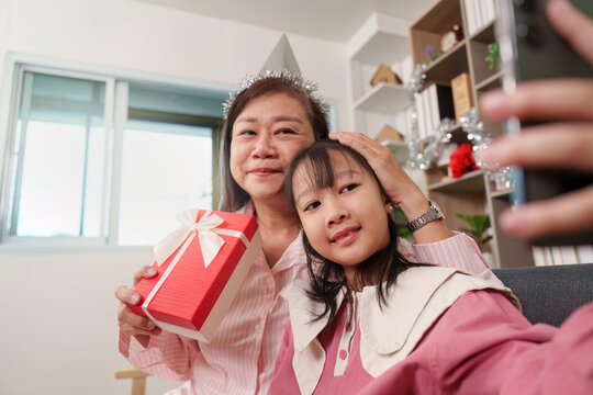 Asian grandmother and granddaughter taking selfie with smartphone while holding gift box at home for birthday celebration