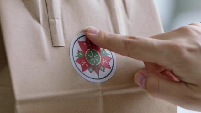 Reaching right hand placing round floral sticker onto brown paper bag front on countertop, securing