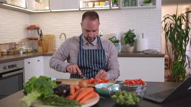 Millennial man watching online cooking tutorial and cutting tomatoes in the kitchen