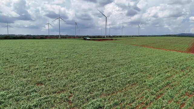 Drone footage captured at low altitude above cultivated farmland in Gia Lai Province, Vietnam. The drone skims over green agricultural fields while wind turbines appear on the distant horizon. The foo