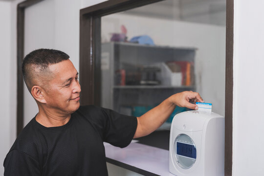A middle aged asian office worker inserts attendance card into a time recorder machine to clock in for work, tracking employee hours and workplace attendance.