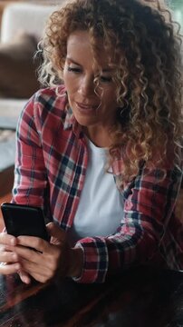 Beautiful woman with curly hair happily smiling and looking at her cellphone while sitting at a table. Online dating and social media application concept for modern adult female people