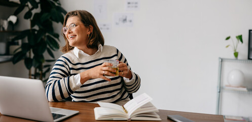 Holding a mug, the woman pauses during laptop work in her home office