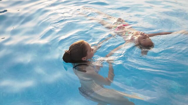 An older sister helps her little sister in a pink swimsuit learn to swim in a pool of clear water