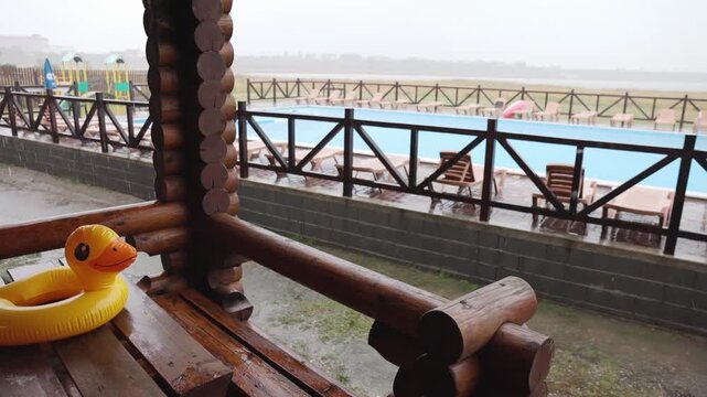 An inflatable duck lies on wooden table in gazebo at recreation center against the backdrop of pool covered with rain