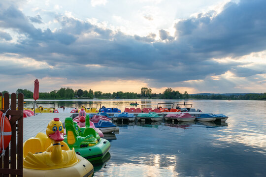 Colorful pedal boats line a floating dock under dramatic evening clouds and sun rays over the lake. Reflections on calm water create a peaceful summer recreation scene.