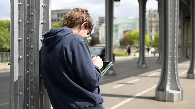 A young man stands outside next to a metal column while holding a tablet. Seen from a three-quarters back view, he uses the device near a city street in Paris.