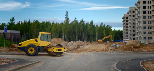 Yellow road roller and excavator on construction site with unfinished building and forest in the background under a clear blue sky © Андрей Знаменский
