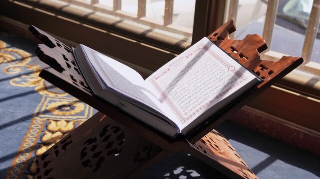 An open Holy Quran rests on a carved wooden stand beside a bright window. Strong daylight casts dramatic shadows across the Arabic text and blue patterned mosque carpet during a peaceful morning.
