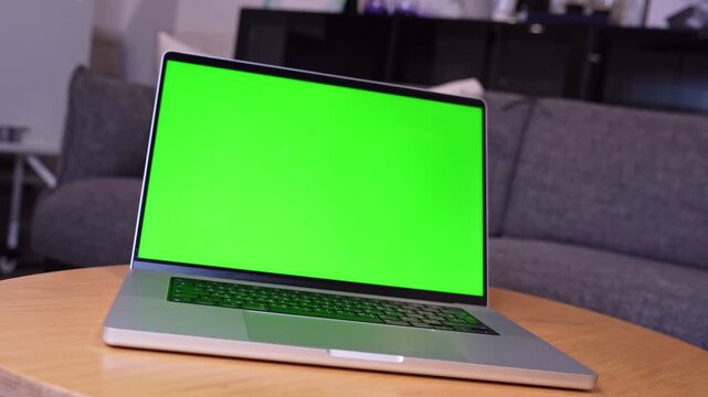 An open silver laptop displaying a bright green screen rests on a round wooden table. The computer sits inside a modern living room with a softly blurred grey sofa in the background during daylight.