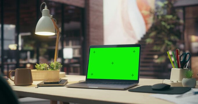 360 Degree Orbital Tracking Shot of Green Screen Chroma Key Laptop on Wooden Desk in Loft Office, Showcasing Blank Mockup Display With Lamp, Coffee Mug and Plants in a Modern Creative Workspace.