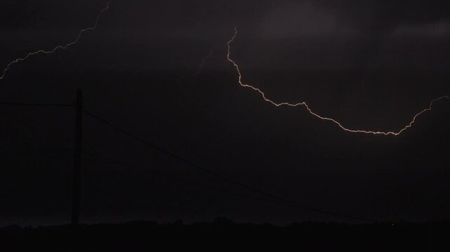 A powerful bolt of lightning illuminates a dark rural landscape with silhouetted power lines, capturing the intense, moody energy of a nighttime storm.