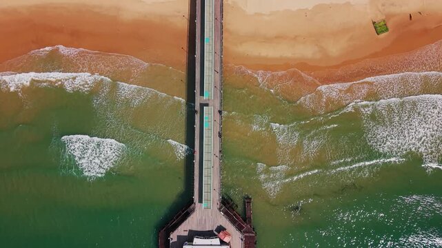 Top down aerial drone footage of Bournemouth Pier crossing over the sandy beach and turquoise sea, with waves breaking along the shoreline below. Useful for themes of UK travel, tourism