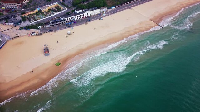 Aerial drone footage of Bournemouth beach showing turquoise sea, white waves, sandy shoreline, promenade, seafront buildings and coastal groynes along the Dorset coast. Useful for themes of UK travel,