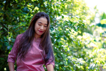 Fototapeta premium A young woman smiles softly beneath the green leaves. Her pink plaid shirt adds a cheerful contrast.