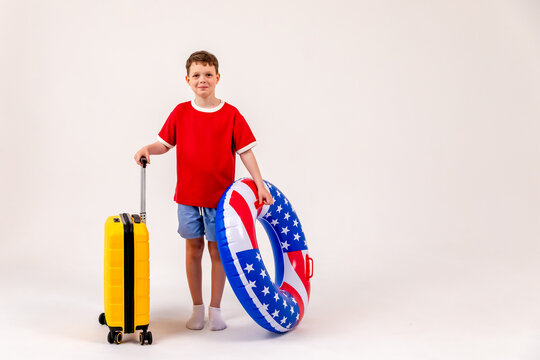 Little boy in red t-shirt and blue shorts holds yellow suitcase in one hand and swimming pool ring with American flag design in other on white background. Summer vacation in USA concept. 4th of July