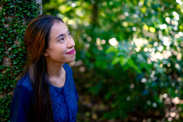 Naklejka premium A young woman smiles warmly beside a tree. The green leaves glow in the soft natural light.