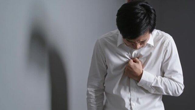 Experiencing chest pain, Asian man doubling over holding chest by gray wall, white shirt and shadow