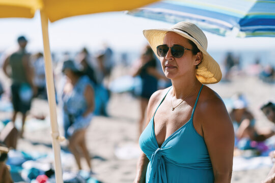 A relaxed woman wearing sunglasses and a hat stands under a beach umbrella, enjoying the sunny day at the beach, portraying leisure and the joy of summer outings.