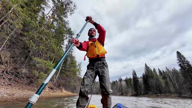 Caucasian paddler on board cruising river in yellow vest and red jacket, navigating rocky shore beneath overcast sky, surrounded by tall pine forest and gentle current, peaceful wilderness