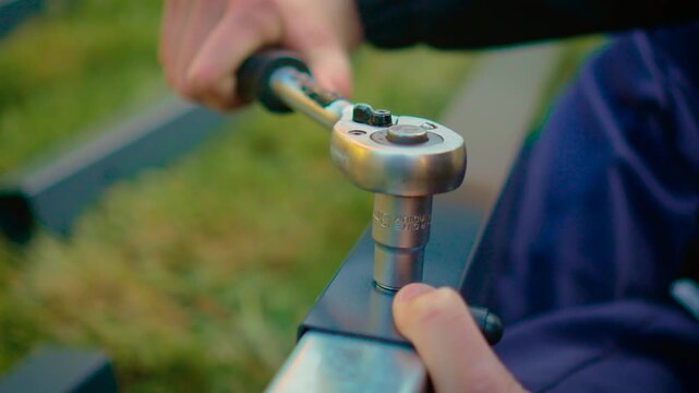 Close angle ratchet wrench engaging bolt on rectangular metal profile in grass area. Repair installation activity with hand tool and structural component alignment. Outdoor construction workflow with