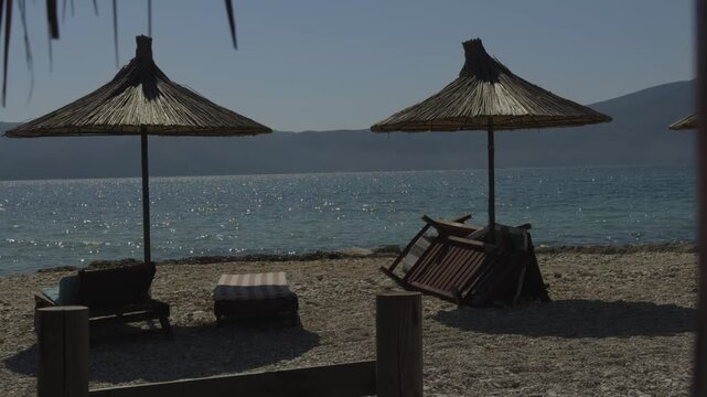 Straw Parasols And Wooden Sunbeds On Pebble Beach Against Sparkling Sea Water. Summer Vacation. Relaxation And Tourism In Mediterranean