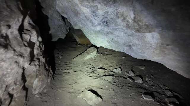 Low-angle interior view of a narrow subterranean passage illuminated by a directional flashlight from the lower-left. A rough vertical limestone wall with visible cracks, mineral
