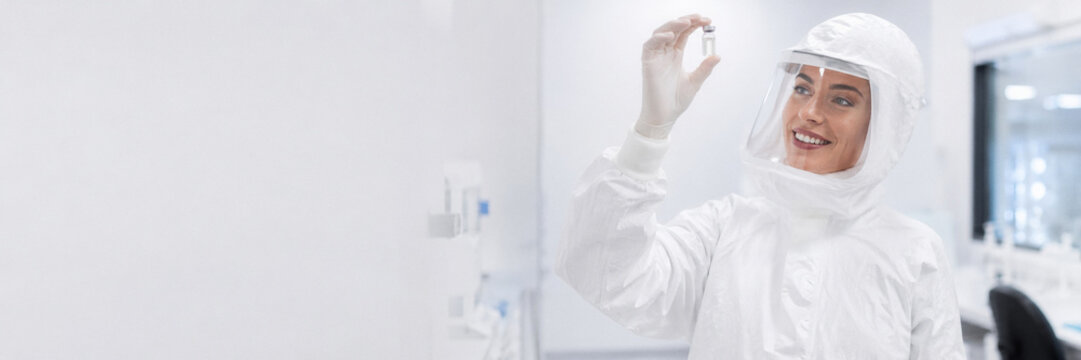 Scientist in a white protective suit and gloves holds a vial while smiling in a modern laboratory. Laboratory research and safety.