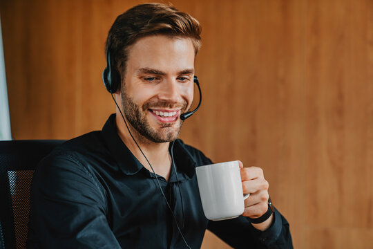 Young man in headset holding coffee cup while working in modern office, professional employee enjoying coffee break during remote work, online communication, corporate lifestyle