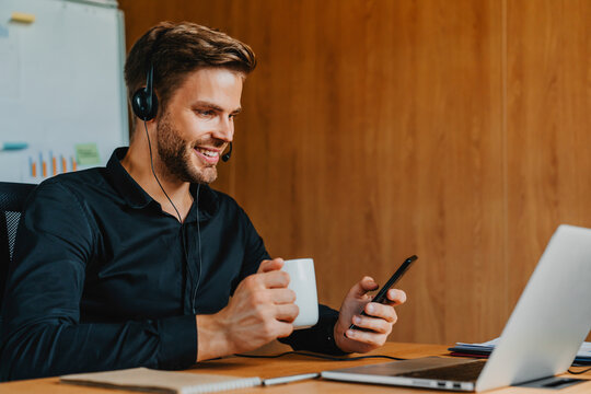 Handsome man in headset drinking coffee and using smartphone while working in modern office, professional employee enjoying coffee break, online communication, multitasking, corporate lifestyle