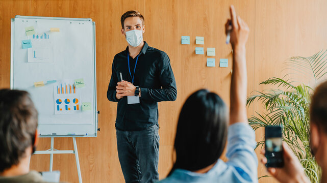 Confident businessman in mask giving corporate presentation to group of coworkers, discussing business project during office seminar, leadership, teamwork collaboration professional training concept