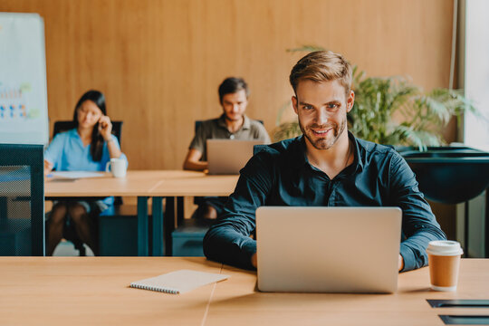 Young man working on laptop in modern startup office with colleagues on background, professional teamwork environment, business communication, corporate workspace, productivity young entrepreneur