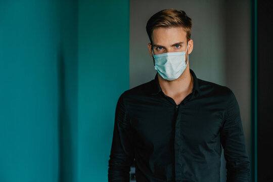 Portrait of young office worker wearing protective face mask looking at camera while standing in modern office, professional businessman during pandemic, healthcare safety and corporate workplace