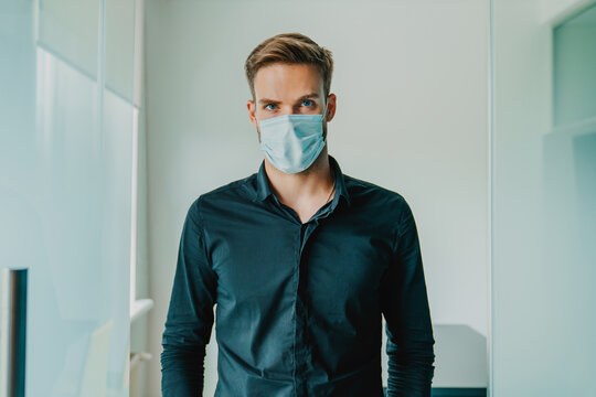 Young man office employee in protective medical mask standing in modern office and looking at camera, business workplace safety, virus prevention, healthcare protection and corporate lifestyle concept