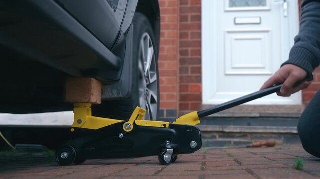 Lifting car with hydraulic floor jack and wooden spacer during vehicle maintenance preparation. Automotive repair concept with manual car raising on driveway for wheel service access. DIY vehicle