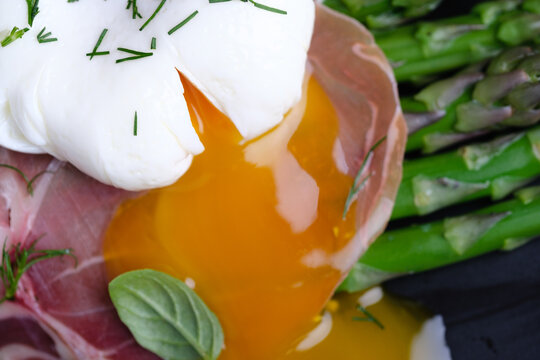 A dish is prepared with poached egg, green asparagus, and thinly sliced ham. The food is arranged on a black plate. This scene is set in a kitchen. Healthy eating concepts.