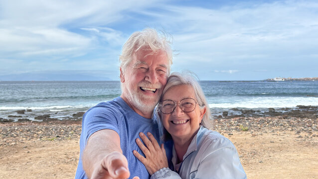 Happy senior couple enjoying retirement vacation at the beach, taking a selfie together, smiling and embracing while appreciating the ocean view and sunny weather