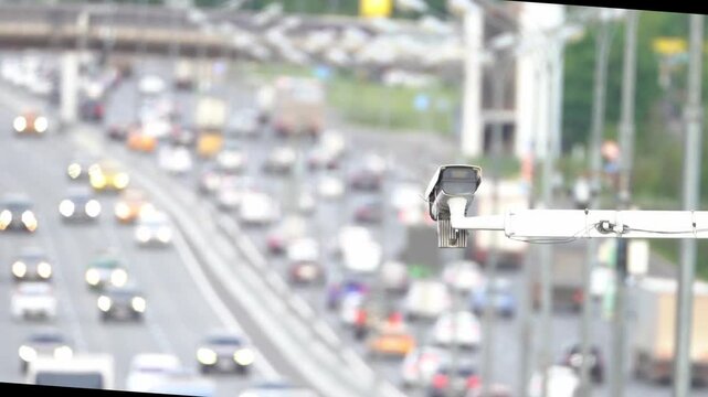 Traffic camera mounted on a pole captures the busy highway. Many cars move steadily on the multi-lane road. Headlights shine bright in the blurred traffic scene